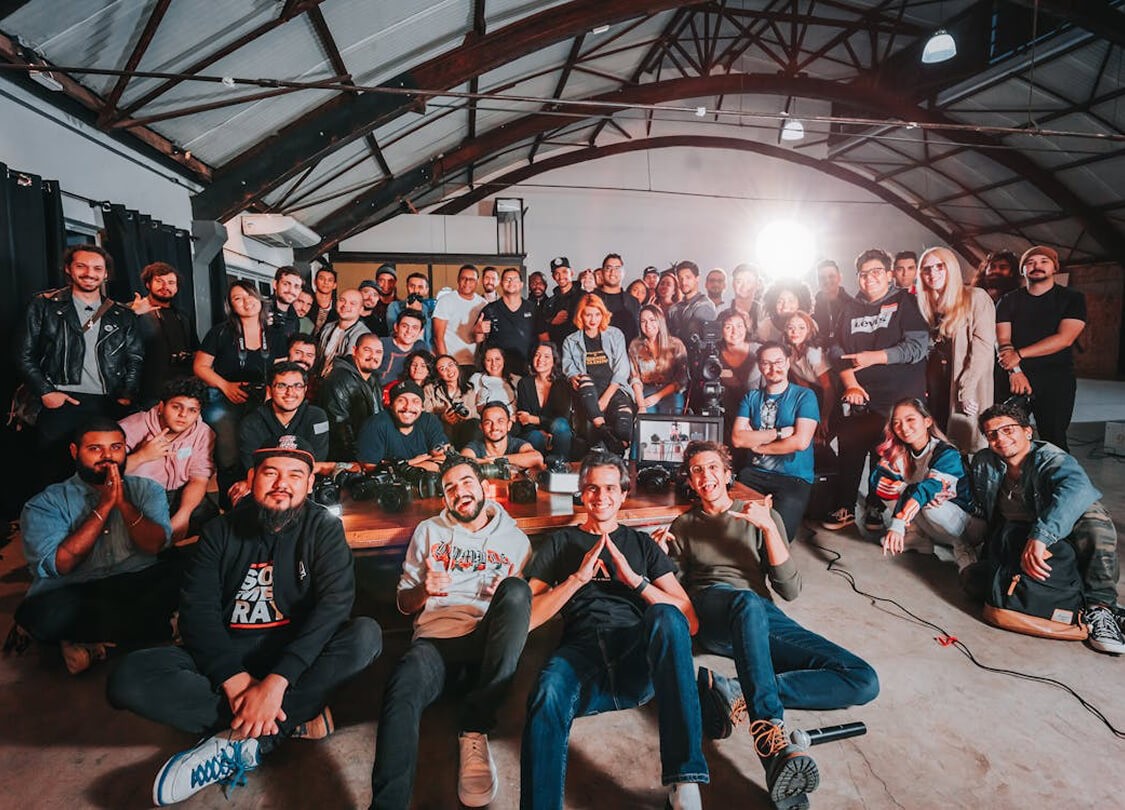 A large group photo of diverse team members gathered in an industrial-style space with arched ceiling.