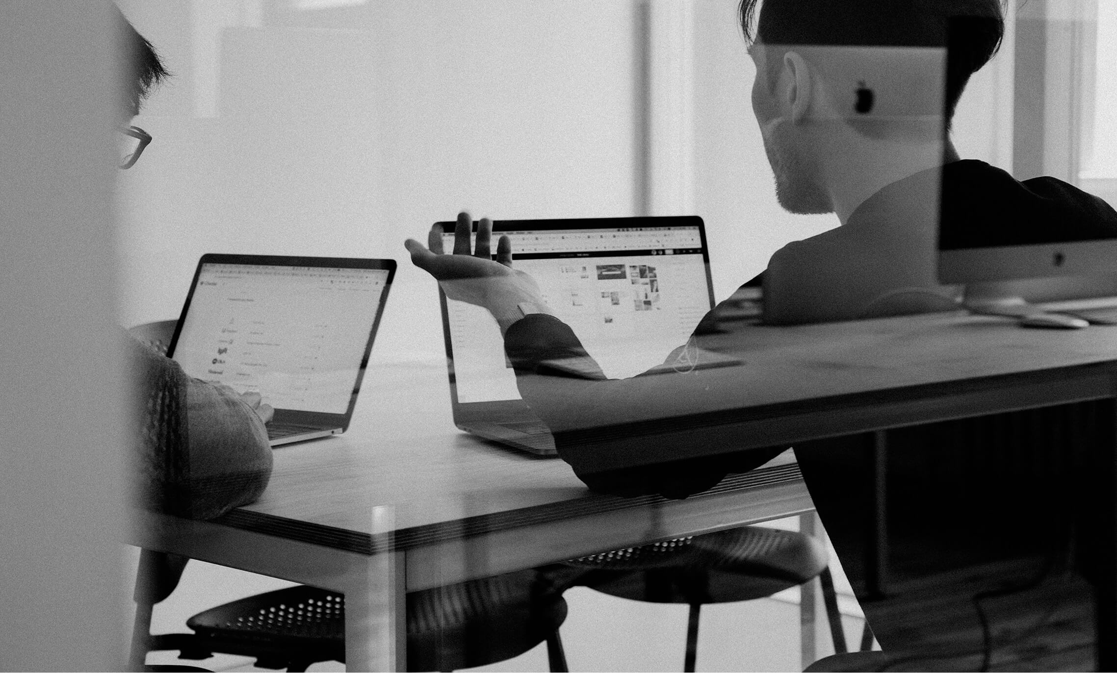 Black and white image of people working at a table with laptops, showing hands gesturing during discussion of digital content displayed on screens, with fabrica® logo in the corner.
