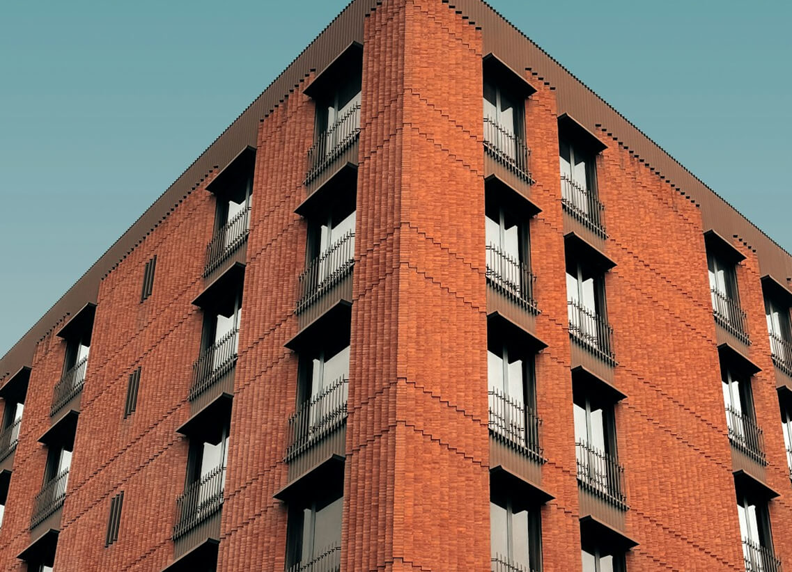Modern red brick apartment or office building viewed from below against a blue sky, showcasing its geometric architecture with evenly spaced windows and balconies.