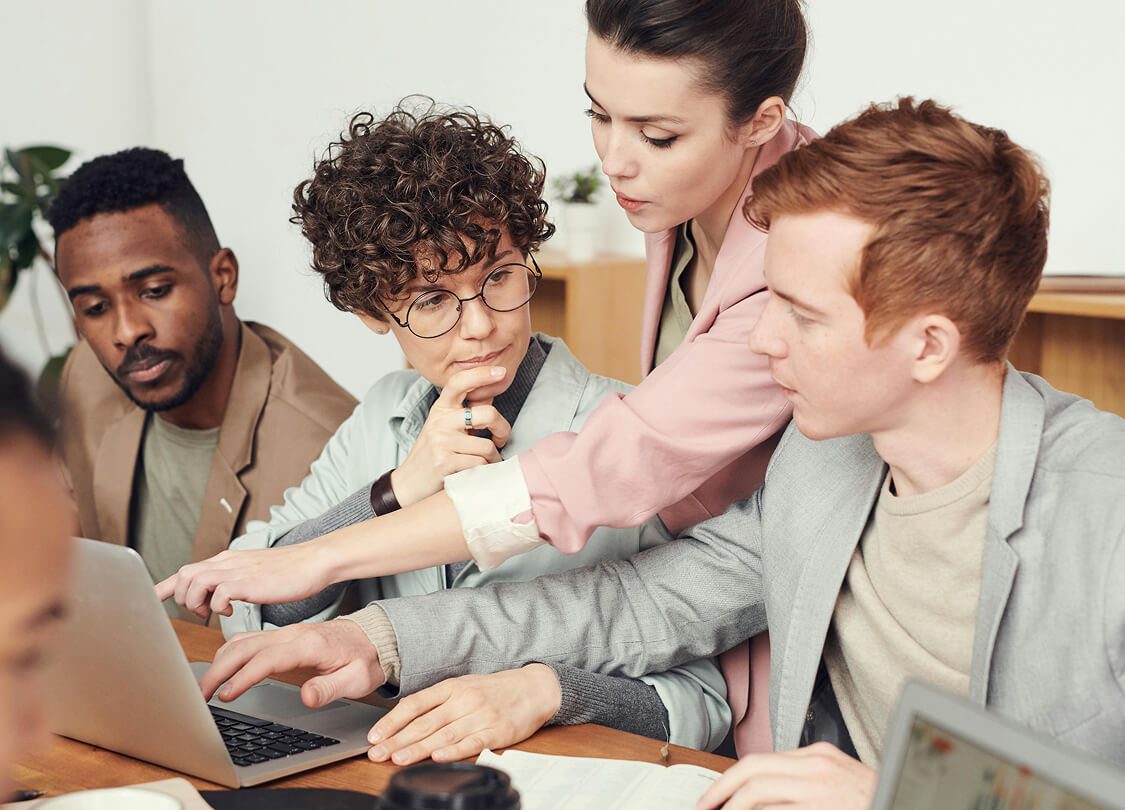 Four diverse professionals collaboratively working on a laptop, with one person pointing at the screen while others observe.